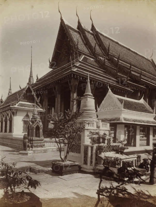 Main building of the temple complex of Wat Pra Keo (Temple of the Emerald Buddha) in Bangkok, album page, photograph, Part of Album with 50 photos of a trip through Southeast Asia., check stamp: 'GR Lambert Singapore', photographer: GR Lambert & Co., Bangkok, c. 1870 - c. 1900, paper, albumen print, height 274 mm x width 209 mm, temple, shrine, Hinduism, Buddhism, Jainism, A large ornate temple complex with steep multi-tiered gabled roofs decorated with upward-curving finials, flanked by smaller roofed structures and spires; a covered colonnade with carved columns and a raised platform; decorative stone and stucco elements including a stupa-like monument, an ornate shrine housing a small statue, and sculpted guardian figures; potted plants and small trees in square planters placed on a paved courtyard; shadowed foreground foliage and stone paving leading toward the buildings.