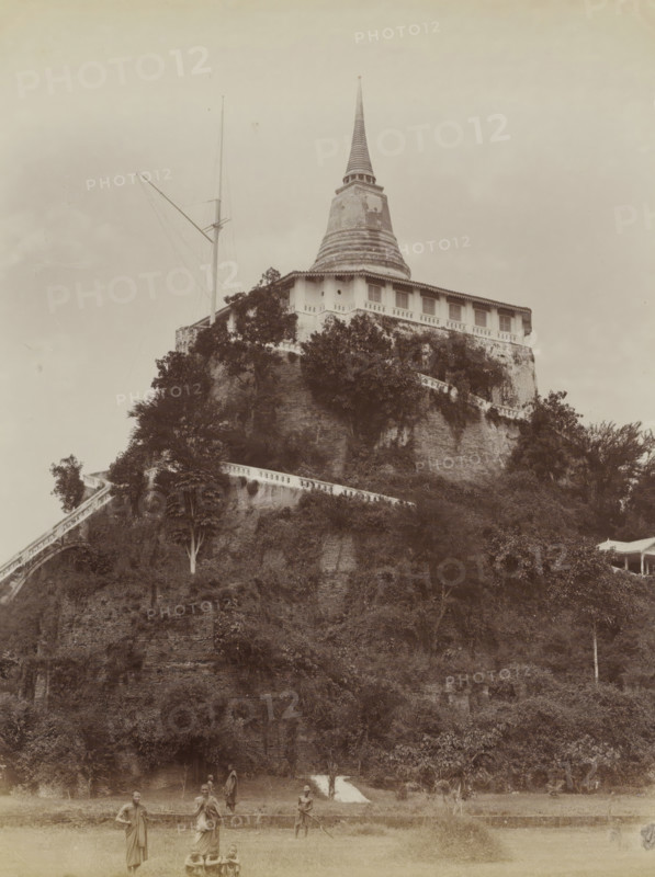 Monks at the foot of the tower of the Wat Saket temple in Bangkok, album page, photograph, Part of Album with 50 photos of a trip through Southeast Asia., check stamp: 'GR Lambert Singapore', photographer: GR Lambert & Co., Bangkok, c. 1870 - c. 1900, paper, albumen print, height 274 mm x width 210 mm, temple, shrine, Hinduism, Buddhism, Jainism, A large tiered structure with a pointed conical top sits atop a steep, vegetation-covered rock face, with a balustraded walkway running along the slope and a flight of steps winding upward. The building near the summit has a covered veranda with evenly spaced window openings and decorative eaves, and a tall slender mast with rigging rises beside it. Dense foliage and trees cling to the hillside, partially obscuring the masonry. In the foreground a grassy open area contains several standing figures wearing draped garments and a small group of seated children. The sky above is mostly clear with a pale, even tone.
