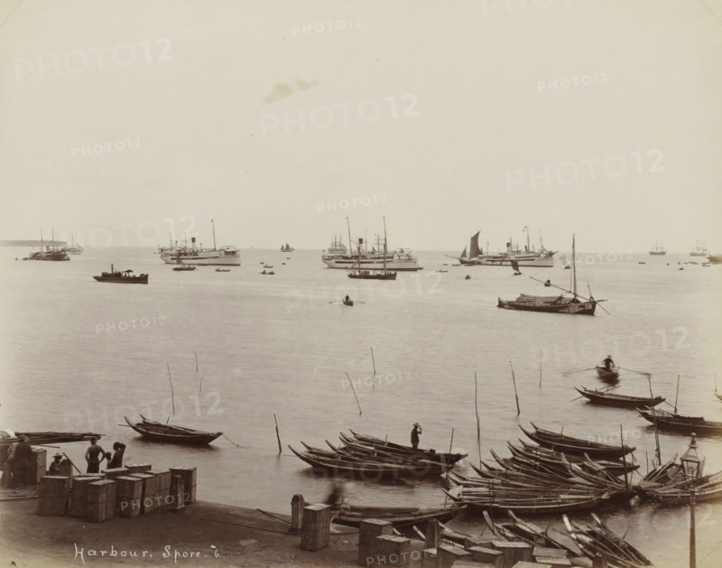 Harbour, S.pore, Ships and rowboats in the harbour of Singapore, album page, photograph, Part of Album with 50 photos of a trip through Southeast Asia., check stamp: 'GR Lambert Singapore', negative number: '6', photographer: GR Lambert & Co., Singapore, c. 1870 - c. 1900, paper, albumen print, height 210 mm x width 268 mm, A wide view of a harbor scene showing calm water with numerous boats and ships at varying distances, small wooden rowboats and narrow longboats clustered near the foreground alongside a stone quay where crates and bollards are visible, several people standing or sitting on the quay and on some boats, a few figures rowing in small boats on the water, a larger sailboat with a dark sail and several steam or motor ships farther out, slender poles rising from the water among the boats, and a pale sky above the horizon.