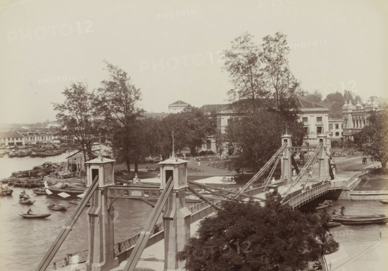 Cavenach Bridge, S'pore, Cavenach Bridge in Singapore, album page, photograph, Part of Album with 50 photos of a trip through Southeast Asia., check stamp: 'GR Lambert Singapore', negative number: '10', photographer: attributed to GR Lambert & Co., Singapore, c. 1870 - c. 1900, GR Lambert & Co., photographer, Attributed to, paper, albumen print, height 209 mm x width 274 mm, bridge in city across river, canal, A sepia-toned scene of a suspension bridge crossing a river, with ornate stone towers and metal cables, several boats moving in the water below, and a riverside area with small wooden structures and stacked timber. On the far bank there are trees with dense foliage partially obscuring large classical-style buildings with columns and tiled roofs. People appear as small figures on the bridge and along pathways near the buildings, and a carriage is visible on a road beside the riverside structures. The sky is pale and empty, and foreground foliage frames the lower part of the image.