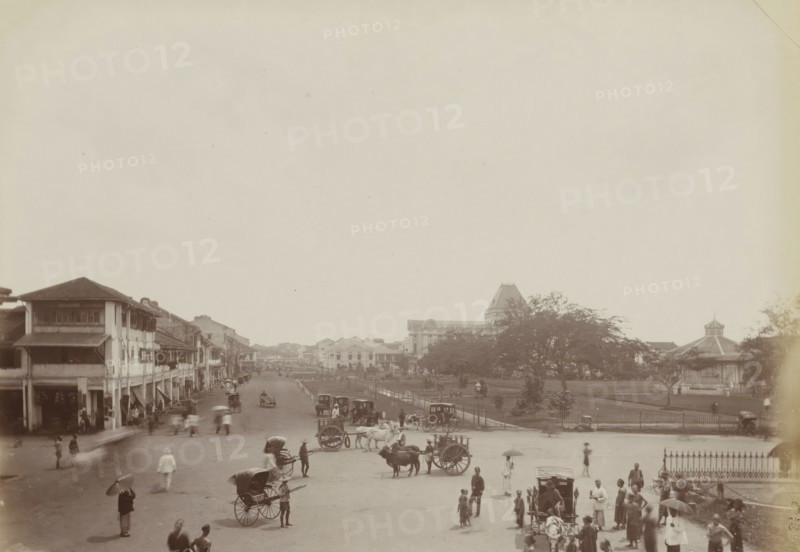 View of a square behind a courthouse in Singapore, View at back of Police Court. S.pore, album page, photograph, Part of Album with 50 photos of a trip through Southeast Asia., check stamp: 'GR Lambert Singapore', negative number: '65', photographer: GR Lambert & Co., Singapore, c. 1870 - c. 1900, paper, albumen print, height 211 mm x width 275 mm, square, circus, etc. (city(-scape) with figures, staffage), A wide street scene with a row of two-story buildings with covered balconies and awnings along the left side, a broad open road filled with various horse-drawn carts and rickshaws, and numerous small groups of people walking or standing across the foreground and midground. On the right side there is a fenced green space with trees and a bandstand with a pointed roof, and behind it larger buildings including one with a tall roof and ornate upper structure. The overall image is in a faded monochrome tone.