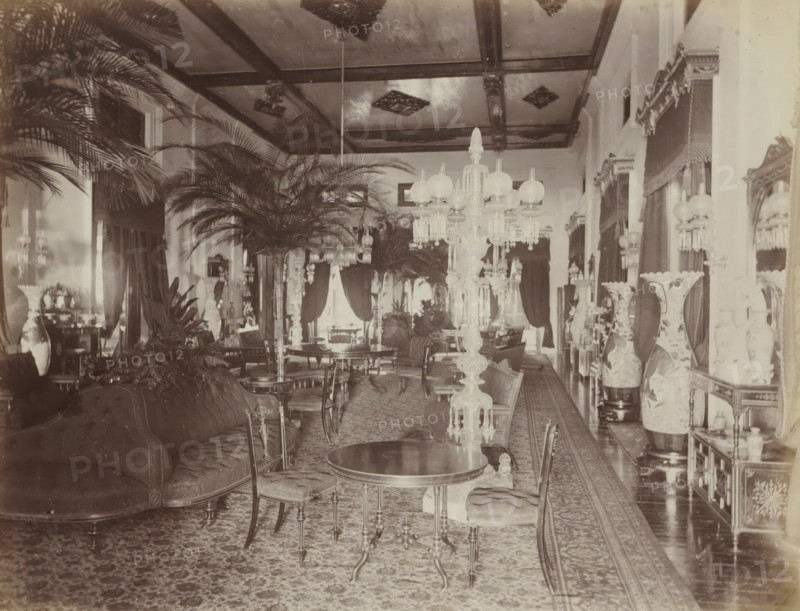 Salon in the palace of the Sultan of Johor, album page, photograph, Part of Album with 50 photos of a trip through Southeast Asia., check stamp: 'GR Lambert Singapore', photographer: GR Lambert & Co., Malaysia, c. 1870 - c. 1900, paper, albumen print, height 211 mm x width 275 mm, drawing-room, 'salon', A long ornate room filled with upholstered sofas and wooden chairs around circular and oval tables, tall decorative floor lamps with glass shades and hanging crystal prisms, large potted palm plants and other foliage arranged along the center and sides, patterned carpets covering the floor, tall vases with painted designs on stands along one side, heavy draped curtains framing tall windows, large mirrors and framed openings on the walls, a coffered ceiling with decorative panels, and various small decorative objects and furniture pieces placed throughout the space.