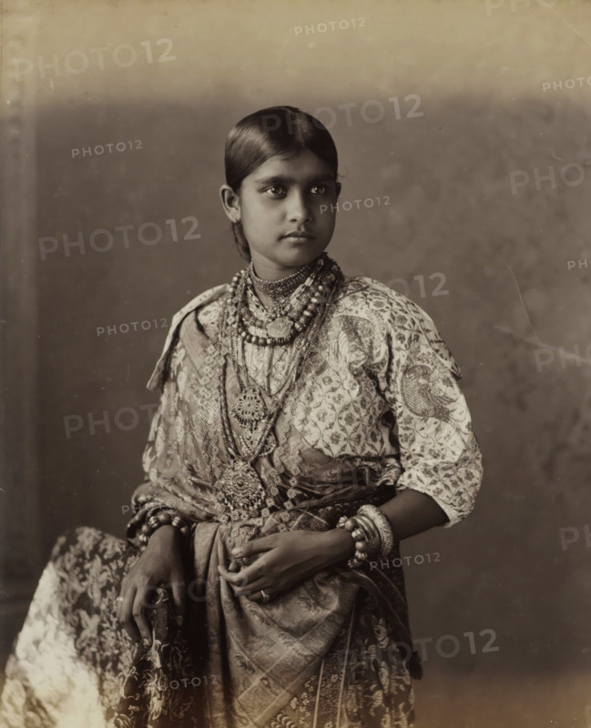 Kandian Lady, Portrait of a young woman from Kandy in Ceylon, album page, photograph, Part of Album with 50 photographs of a trip through Southeast Asia., photographer: Charles T. Scowen, Sri Lanka, c. 1875 - c. 1880, South Asia, paper, albumen print, height 278 mm x width 216 mm, adolescent, young woman, maiden, A young person with neatly parted dark hair and a calm expression is shown from the waist up wearing patterned clothing and multiple layered necklaces and pendants, bracelets and bangles on both wrists, and a patterned wrap draped across the body, with hands resting in front and a plain backdrop behind.