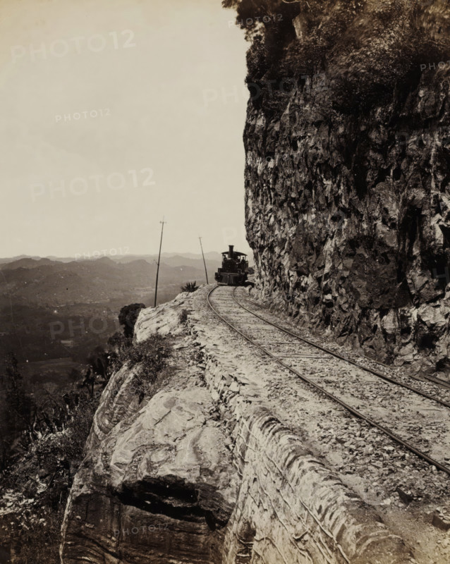 Train runs along a precipice at Sensation Rock in Ceylon, Sensation Rock. Colombo and Kandy Line., album page, photograph, Part of Album with 50 photos of a trip through Southeast Asia., photographer: Charles T. Scowen & Co., Sri Lanka, c. 1876 - c. 1885, South Asia, paper, albumen print, height 277 mm x width 217 mm, railway, train, mountains, A narrow railway track runs along a rocky cliff edge with a steam locomotive visible ahead on the track, partially framed by the cliff face on the right; the cliff shows jagged rock textures and vegetation at the top, while the left side opens to a distant hilly landscape under a pale sky and a couple of leaning telegraph poles rise near the track.