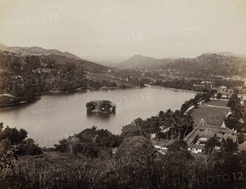 View of Lake Kandy in Ceylon, album page, photograph, Part of Album with 50 photos of a trip through Southeast Asia., stamp: 'Fabrikmarke', photographer: Charles T. Scowen & Co., Kandy (former nation), c. 1875 - c. 1880, South Asia, paper, albumen print, height 215 mm x width 275 mm, lake, A wide sepia-toned landscape showing a large calm lake with a small vegetated island near its center, tree-covered hills rising around the lake, a shoreline with scattered buildings and clusters of trees, a long low bridge or causeway crossing part of the water, swaths of lawn and formal grounds with rows of tall palms near a complex of tiled-roof buildings in the foreground, dense foliage and grassy foreground slopes framing the bottom of the scene, and distant layered hills fading toward the horizon under a pale sky.
