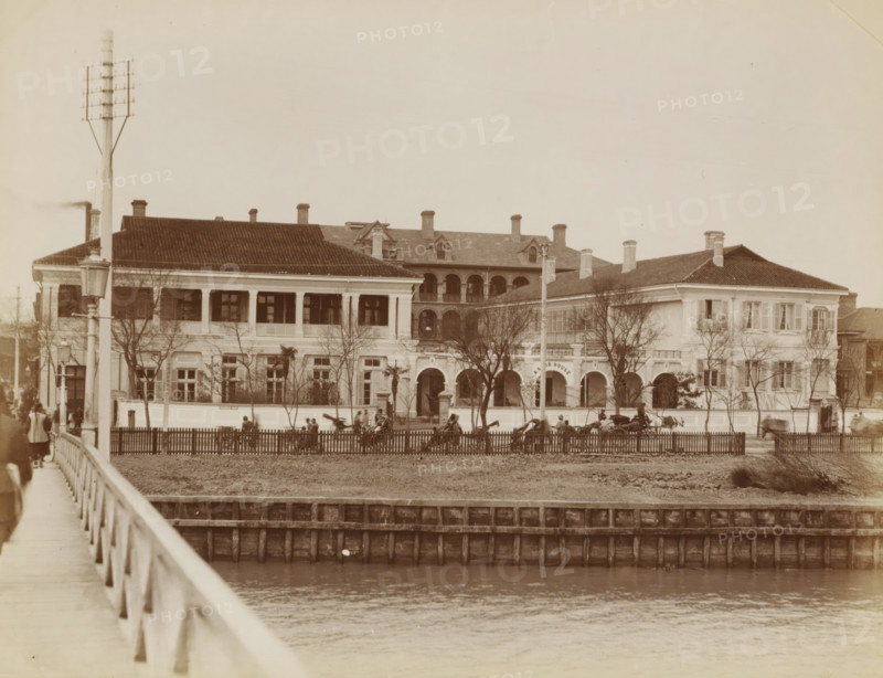Exterior of the Astor House Hotel in Shanghai, album page, photograph, Part of Album with 50 photos of a trip through Southeast Asia., Shanghai, in or after 1859 - c. 1900, photographer, paper, albumen print, height 204 mm x width 274 mm, hotel, hostelry, inn, A sepia-toned photograph shows a large two-story building with a tiled roof and multiple chimneys, featuring an open upper veranda with columns and an arched central entrance flanked by windows and smaller arches; leafless trees stand in front of the building and a fenced lawn separates it from a body of water in the foreground, with a wooden bridge and railing extending along the left side of the image; several horse-drawn carriages and groups of people are visible near the fence and entrance, and a tall utility pole with crossarms stands near the left front of the building.