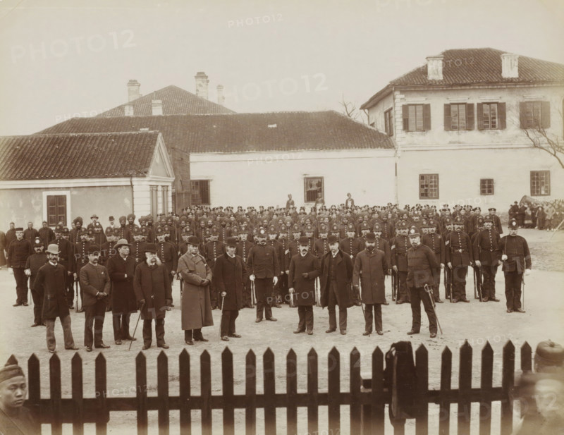 Group portrait of Western policemen of the Shanghai International Concession, album page, photograph, Part of Album with 50 photos of a trip through Southeast Asia., Shanghai, c. 1880 - c. 1910, photographer, paper, albumen print, height 212 mm x width 271 mm, policeman, constable, A large group of men arranged in rows stands in front of a cluster of light-colored buildings with tiled roofs and shuttered windows; many of the men wear dark uniforms and peaked or bowler hats, some hold canes or swords, and a few wear long overcoats and facial hair; behind them a few taller figures stand near windows and doorways, and a wooden picket fence runs across the foreground.
