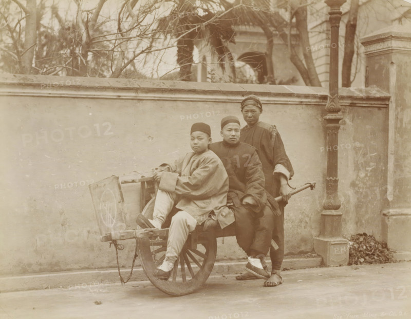Three Chinese people in Shanghai use a wheelbarrow as a means of transport, album page, photograph, Part of Album with 50 photos of a trip through Southeast Asia., address: '42 Nanking Road', photographer: Sze Yuen Ming & Co, Shanghai, c. 1860 - c. 1900, paper, albumen print, height 212 mm x width 270 mm, wheelbarrow, Three people are positioned around a wooden cart with a large spoked wheel in front of a stone wall; two sit on the cart while one stands behind holding the cart's handle. The seated individuals wear long, loose garments and round caps, and the standing person wears a long dark robe with a wrapped head covering and sandals. The cart shows visible wood grain and rope attachments, and a circular pattern is faintly visible on the cart's side. Behind them, a wall topped with a ledge runs horizontally across the scene, with leafless tree branches and parts of a building visible above the wall. A tall ornate lamppost or column stands to the right against the wall, and a small p...