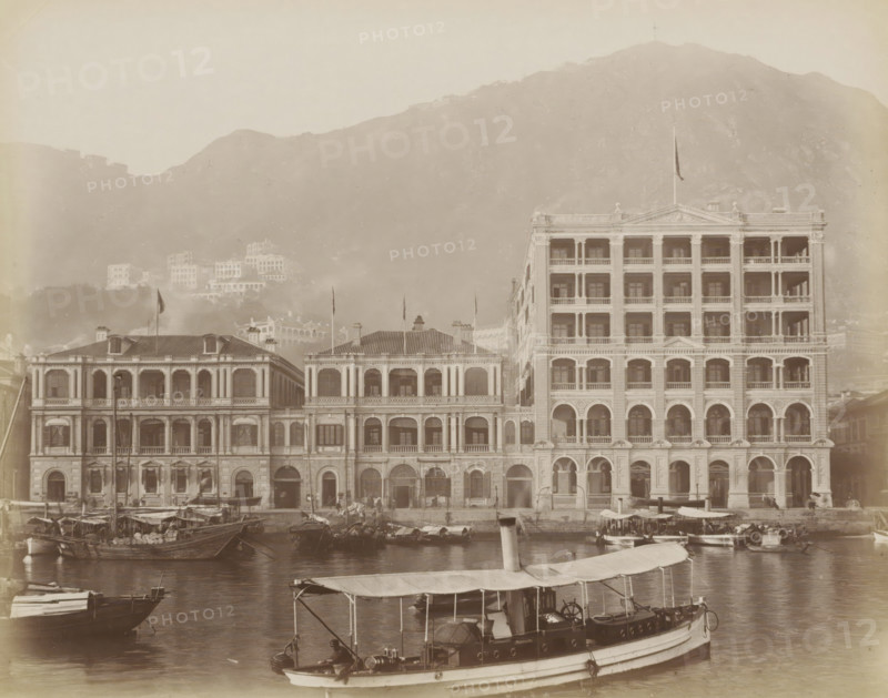 View of the Hong Kong Hotel in Hong Kong, album page, photograph, Part of Album with 50 photos of a trip through Southeast Asia., Hong Kong, c. 1860 - c. 1900, photographer, paper, albumen print, height 223 mm x width 284 mm, hotel, hostelry, inn, A waterfront scene showing a row of ornate multi-story buildings with arched ground-floor openings, columns, balconies, and decorative facades, with a taller building on the right and lower connected buildings to the left; small boats and sampans are moored in the water in the foreground, one larger covered boat with a canopy and visible equipment is closest to the viewer; hills rise behind the buildings, with additional structures partially visible on the slopes and a cross-shaped structure near the hilltop; the overall image has a sepia tone and a hazy atmosphere.