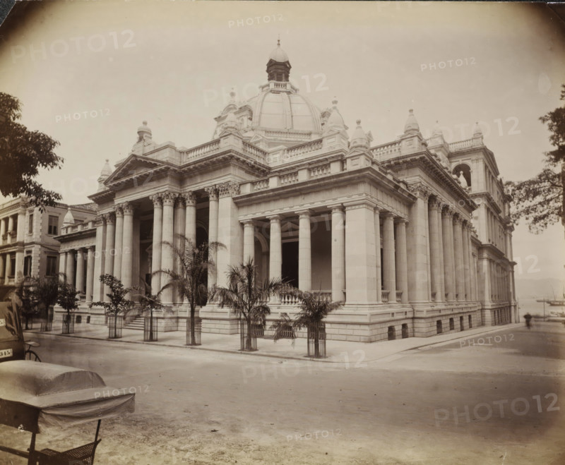 Exterior of the Hong Kong-Shanghai Bank in Hong Kong, album page, photograph, Part of Album with 50 photographs of a trip through Southeast Asia., Hong Kong, c. 1860 - c. 1900, photographer, paper, albumen print, height 233 mm x width 288 mm, exterior, representation of a building, A large neoclassical stone building with a central dome and a cupola rises above a colonnaded facade, showing tall fluted columns with ornate capitals supporting a heavy entablature and balustraded roofline, decorative pediments and sculptural urns along the roof, and an open arched entrance beneath the portico; young palm trees in metal guards line the sidewalk in front of the building, parts of adjacent classical buildings are visible at the sides, a paved street runs in front with a covered bench or cart and a small section of a chair in the foreground, and leafy tree branches frame the left and right edges of the scene.