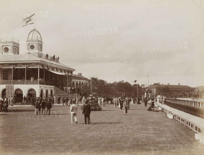 View of the horse racetrack and grandstand of Shanghai, album page, photograph, Part of Album with 50 photos of a trip through Southeast Asia., Shanghai, c. 1890 - c. 1900, photographer, paper, albumen print, height 215 mm x width 277 mm, horse-racing, race-track, A large two-story building with arched openings on the ground level and a covered upper veranda occupies the left side, topped by a tower with a round window and an open dome structure bearing a flag, with people visible on the veranda and along a railing on the roof; in front of the building a wide open lawn stretches toward the foreground where groups of men and women in period clothing are standing, walking, and gathering, some wearing hats and some holding umbrellas, with floral plantings and a raised pathway or promenade flanked by a balustrade on the right leading toward distant low buildings and trees under a broad cloudy sky.
