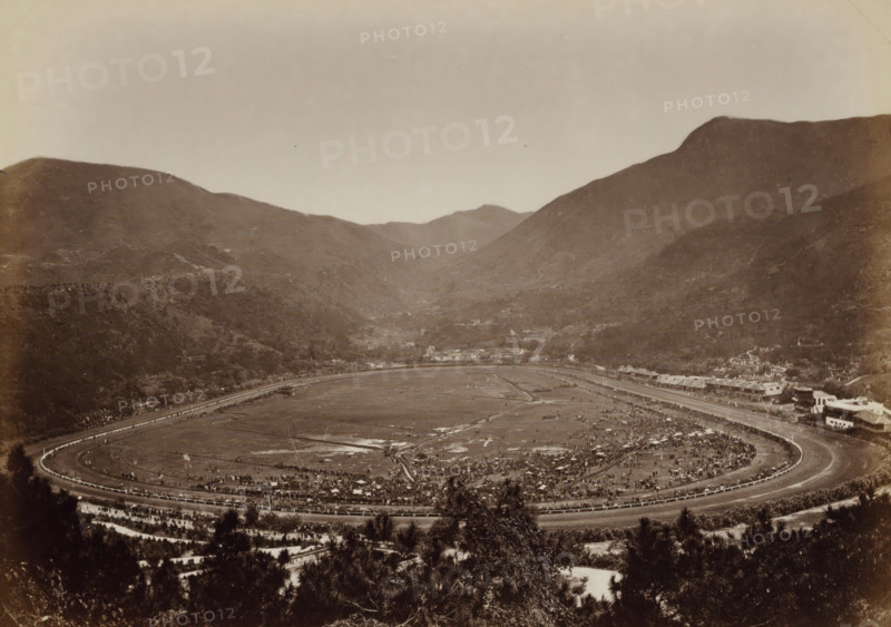 View of Happy Valley, the Hong Kong horse racetrack, album page, photograph, Part of Album with 50 photos of a trip through Southeast Asia., Hong Kong, c. 1860 - c. 1900, photographer, paper, albumen print, height 207 mm x width 291 mm, horse-racing, race-track, A wide, oval racetrack sits in the foreground surrounded by a low fence, with crowds gathered along its inner and outer edges and scattered tents and small structures on the infield; beyond the track a narrow road and a line of low buildings run along the base of a valley, and the scene is framed by steep, tree-covered hills rising on both sides into higher, rounded mountain ridges under a pale sky.