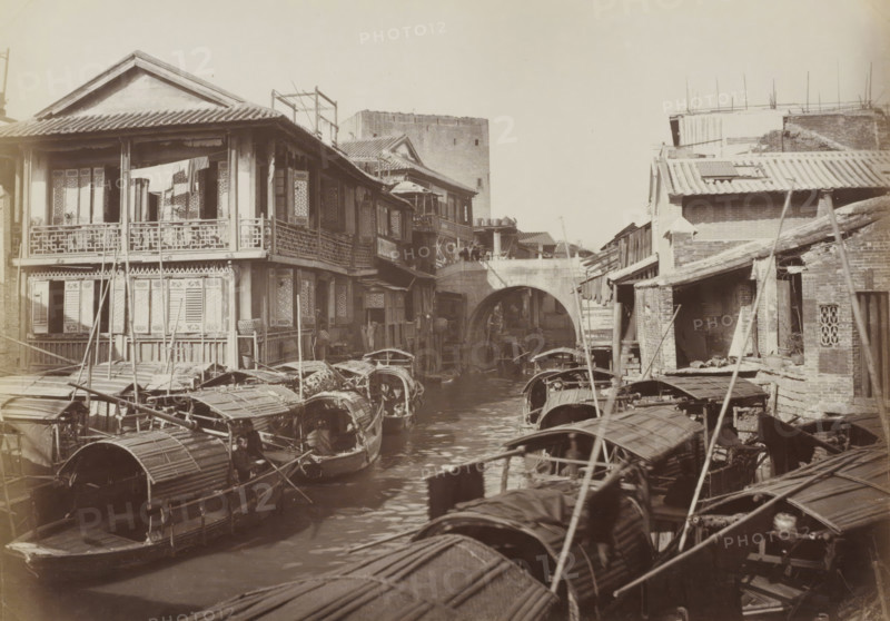 Boats and houses in Canton, album page, photograph, Part of Album with 50 photos of a trip through Southeast Asia., Canton (city), c. 1860 - c. 1900, photographer, paper, albumen print, height 205 mm x width 291 mm, harbour, A narrow waterway crowded with small covered wooden boats stretches toward a stone arched bridge, flanked on both sides by multilevel riverside buildings with tiled roofs and wooden balconies; the left bank shows a larger building with decorative railing and open windows with fabric hanging on the upper balcony, while the right bank has smaller brick and wooden structures with sloped roofs and exposed beams; boats with curved canopies and long poles fill the foreground and middle distance on the water, and a tall rectangular masonry tower rises behind the buildings in the background.