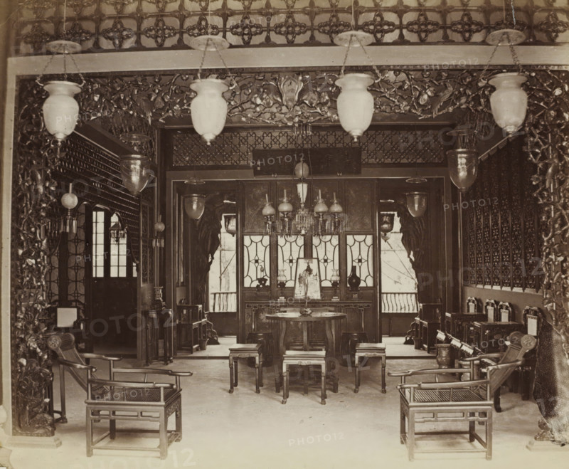 Interior of a Chinese club in Guangzhou, album page, photograph, Part of Album with 50 photos of a trip through Southeast Asia., Guangzhou (city), c. 1860 - c. 1900, photographer, paper, albumen print, height 228 mm x width 275 mm, club, meeting place, An interior room with ornate carved woodwork framing the entrance and decorative patterned screens along the back wall, suspended light fixtures with glass shades hanging from the ceiling, a central round table with stools around it and a chandelier above, pairs of wooden armchairs with open lattice seats positioned near the foreground, additional small tables and cabinets arranged along the sides, carved decorative panels and hanging lanterns visible throughout, and windows with patterned panes letting light into the space.