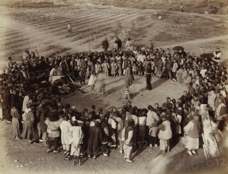 Chinese people in a circle at an outdoor board game, album page, photograph, Part of Album with 50 photos of a trip through Southeast Asia., title: 'Board game', number: '550', China, c. 1860 - c. 1900, photographer, paper, albumen print, height 214 mm x width 278 mm, sports, games and physical performances, A large crowd of people stands and sits outdoors arranged in a rough circle around a central open area where a few figures stand and one person is seated on the ground with nearby bowls and simple objects, many people wear layered garments and head coverings, some hold umbrellas, and several small children are visible among the adults; in the background there are terraced earth beds and a sloped grassy area with a few scattered individuals and a small dog, and the scene is captured in a sepia-toned photograph.
