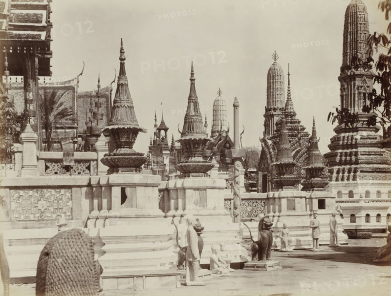 Wat Pra Keo, B.kok, Temple complex of Wat Pra Keo (Temple of the Emerald Buddha) in Bangkok, album page, photograph, Part of Album with 50 photos of a trip through Southeast Asia., check stamp: 'GR Lambert Singapore', photographer: GR Lambert & Co., Bangkok, c. 1870 - c. 1900, paper, albumen print, height 209 mm x width 273 mm, temple, shrine, Hinduism, Buddhism, Jainism, Stone terraces and carved balustrades fill the foreground, topped by ornate bell-shaped and spire-like structures with layered decorative bands and pointed finials. A series of tall, cylindrical towers and sculpted prangs rise in the midground, each covered in intricate relief and patterned surfaces. Several statues and sculpted figures stand or kneel along the steps and platforms, including human forms and animal-like guardians with curved tails and seated postures. Ornamental rooflines with upturned eaves and hanging decorative elements are visible at the left, while leafy branches frame part of the scene at the rig...
