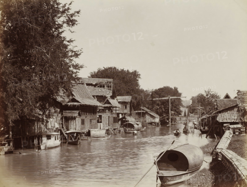 Village by a water in Siam, album page, photograph, Part of Album with 50 photos of a trip through Southeast Asia., check stamp: 'GR Lambert Singapore', inscription: 'Siamese Village at Klang San', photographer: GR Lambert & Co., Thailand, c. 1870 - c. 1900, paper, albumen print, height 210 mm x width 274 mm, river bank, A riverside scene with wooden stilt houses and thatched roofs lining both sides of a narrow waterway, small boats and covered canoes moored along the banks, a larger covered boat in the foreground tied to the shore, narrow wooden walkways and ladders connecting houses to the water, leafy trees rising behind the houses, a few wooden poles and simple bridges spanning the canal, reflections of the buildings and boats visible on the water surface, and an open sky occupying the upper part of the image.