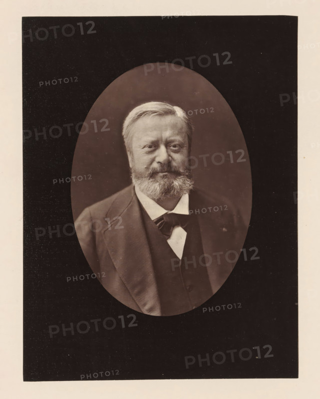 Edmond About .Ne a Dieuze (Meurthe), le 14 fevrier 1828, Portrait of Edmond About, photomechanical print, inscription: '126, boul. Magenta.-Paris.', photographer: Félix Nadar, cliché maker: Goupil & Cie., c. 1876 - in or before 1879, paper, Woodburytype, Woodburytypepie, height 201 mm x width 153 mm, Person, Edmond About, portrait of a writer, A sepia-toned oval portrait of a middle-aged man with light hair and a full beard and mustache set against a dark background, showing his head and upper torso dressed in a dark suit jacket and waistcoat over a light shirt with a visible bow or necktie, all framed by a rectangular dark mat with a light border.