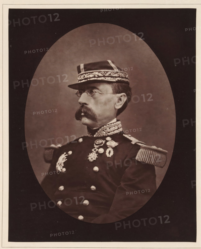 Louis-Leon-Cesar Faidherbe. Ne a Lille, le 3 juin 1818, Portrait of Louis Léon César Faidherbe, page, photomechanical print, inscription: '126, boul. Magenta.-Paris.', photographer: Étienne Carjat, cliché maker: Goupil & Cie., c. 1876 - c. 1879, paper, Woodburytype, Woodburytypie, height 230 mm x width 186 mm, Person, Louis Léon César Faidherbe, commander-in-chief, general, marshal, (military) uniforms, A sepia-toned oval portrait of a man in a formal military uniform shown in profile, facing left, wearing a peaked cap with ornate embroidered band and stripes, small round spectacles, a large handlebar mustache, an embroidered high collar, epaulettes with fringe on the shoulders, and a row of decorative medals and ornate buttons on the chest; the portrait is set against a plain backdrop and surrounded by a dark mat with a light border.