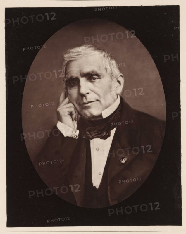 Portrait of Eugène Scribe, Eugene Scribe. Ne a Paris, le 25 decembre, decede le 20 fevrier 1861, page, photomechanical print, inscription: '126, boul. Magenta.-Paris.', photographer: Félix Nadar, cliché maker: Goupil & Cie., c. 1850 - in or before 1861, paper, Woodburytype, Woodburytypepie, height 228 mm x width 182 mm, Person, Eugène Scribe, portrait of a writer, An oval-framed sepia portrait of an older man shown from the chest up, with light-colored hair and sideburns, wearing a dark suit jacket, waistcoat, white shirt, and a dark cravat; he rests his head on his hand with fingers touching his cheek, looking slightly forward with a neutral expression, and a small decorative pin is visible on his jacket lapel.