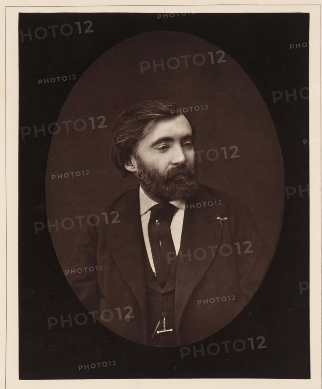 De Bornier. Ne a Lunel, en 1825, Portrait of Henri de Bornier, page, photomechanical print, inscription: '126, boul. Magenta.-Paris.', photographer: Ferdinand Mulnier, cliché maker: Goupil & Cie., c. 1876 - in or before 1879, paper, Woodburytype, Woodburytypepie, height 229 mm x width 182 mm, Person, Henri de Bornier, writer, poet, author, A sepia-toned oval portrait of a man from the chest up, showing a head with dark, combed hair and a full beard and mustache, a contemplative gaze directed slightly to the side, wearing a dark suit jacket over a waistcoat and a white shirt with a dark tie, a small pin or flower on the jacket lapel, and a watch chain visible at the waist, set against a darker rectangular background with a light border.