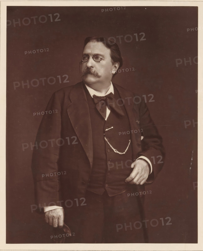 H. Giacomelli. Ne a Paris, le 1er avril 1822, Portrait of Hector Giacomelli, page, photomechanical print, inscription: '126, boul. Magenta.-Paris.', photographer: Ferdinand Mulnier, cliché maker: Goupil & Cie., c. 1876 - in or before 1879, paper, Woodburytype, Woodburytypepie, height 229 mm x width 185 mm, Person, Hector Giacomelli, painting, drawing and the graphic arts, A sepia-toned portrait of a middle-aged man shown from the hips up wearing a dark three-piece suit, a large bow tie, and a pocket watch chain across his vest; he has a full mustache and short, combed hair, and he is wearing pince-nez glasses with a chain; his left hand rests near his waist while his right hand holds a folded hat, and he is looking slightly to his left against a plain dark background.