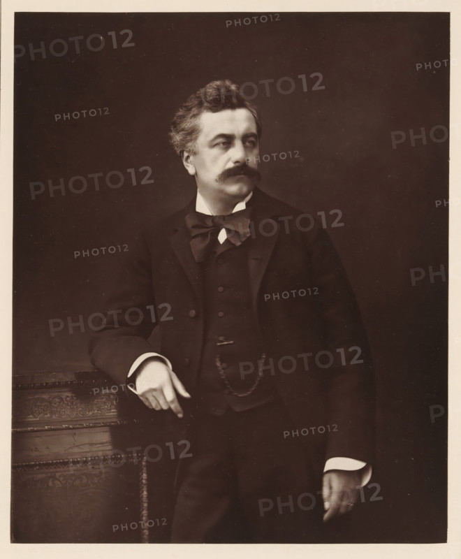 Portrait of Louis Andrieux, L. Andrieux. Ne a Trevoux, le 23 fevrier 1840, page, photomechanical print, inscription: '126, boul. Magenta.-Paris.', photographer: Ferdinand Mulnier, cliché maker: Goupil & Cie., c. 1876 - in or before 1879, paper, Woodburytype, height 225 mm x width 185 mm, Person, Louis Andrieux, politician, eg party leader, A sepia portrait of a man from the waist up wearing a dark three-piece suit with a bow tied at his collar and a visible chain draped across his vest; he has wavy hair, a thick mustache, and is looking slightly to his right; his left arm rests on an ornately carved wooden surface while his right hand hangs by his side showing a ring on a finger; the background is plain and dark.