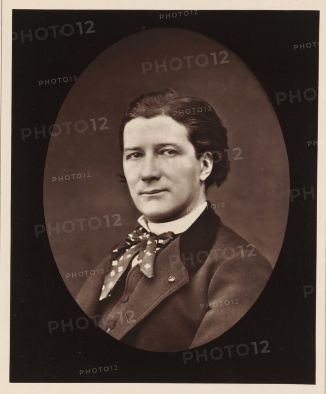 Victorien Sardou. Ne a Paris, le 7 septembre 1831, Portrait of Victorien Sardou, page, photomechanical print, inscription: '126, boul. Magenta.-Paris.', photographer: Ferdinand Mulnier, cliché maker: Goupil & Cie., c. 1876 - in or before 1879, paper, Woodburytype, Woodburytypepie, height 226 mm x width 185 mm, Person, Victorien Sardou, portrait of a writer, A sepia portrait of a person shown from the chest up inside an oval vignette, with dark background surrounding the oval; the person has neatly combed dark hair, a smooth complexion, and a calm facial expression with eyes looking slightly to the side, wearing a high collared shirt, a patterned neckerchief tied in a loose knot, and a dark tailored jacket with a lapel and visible buttons.