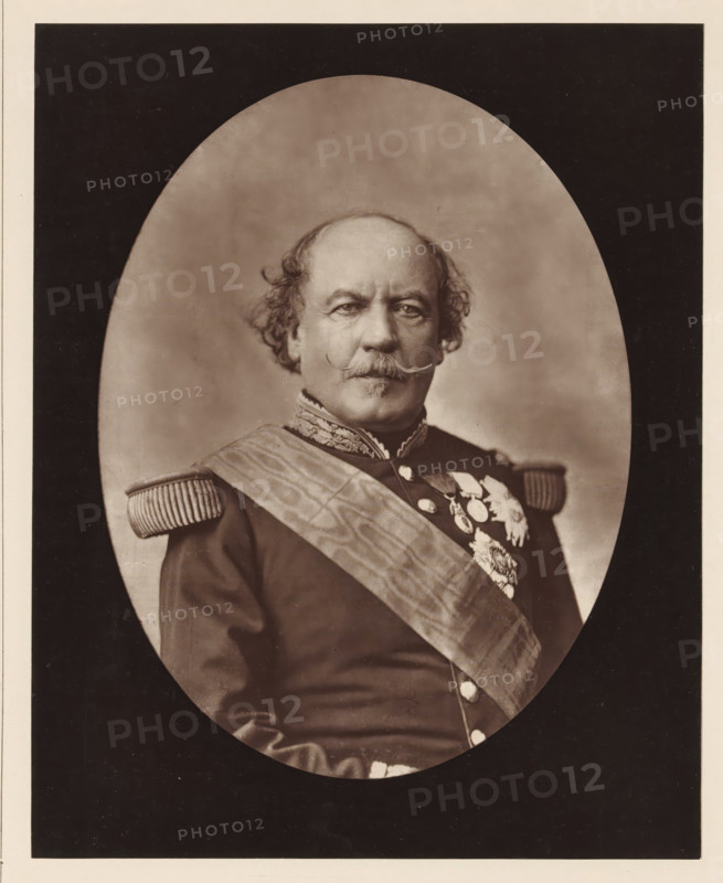 Portrait of François Certain Canrobert, Canrobert. Ne a Saint-Cere (Lot), in June 1809, page, photomechanical print, inscription: '126, boul. Magenta.-Paris.', photographer: Augustin Aimé Joseph Lejeune, cliché maker: Goupil & Cie., c. 1876 - in or before 1879, paper, Woodburytype, Woodburytypepie, height 226 mm x width 181 mm, Person, François Certain de Canrobert, commander-in-chief, general, marshal, A sepia-toned oval portrait of a man shown from the torso up, set against a darker rectangular background. He has a balding head with curly hair at the sides and a prominent curled mustache. He wears a dark military-style uniform with ornate embroidered collar and fringed shoulder epaulettes, a broad sash crossing his chest, and a cluster of decorative medals and badges on his left chest. His expression is neutral and he faces forward.