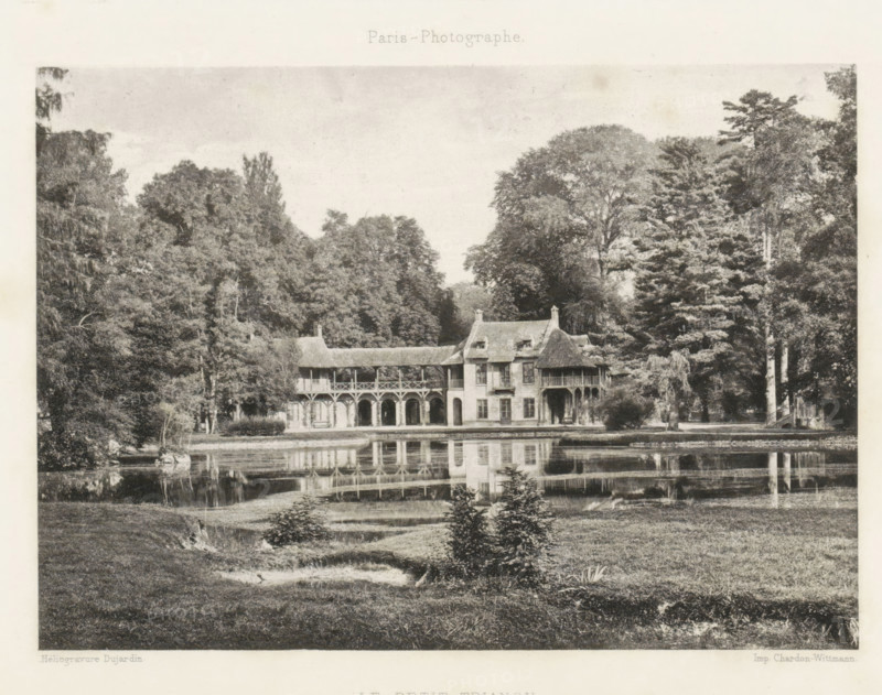 View of Le Hameau de la Reine at Versailles, Le Petit Trianon, page, photomechanical print, Versailles, cliché maker: Dujardin, France, printer (person): Chardon-Wittmann, c. 1881 - in or before 1891, photographer, printer (person), paper, photoaquatint, height 122 mm x width 170 mm, Print on endpaper., farm (building), village, architectural folly, Le Hameau de la Reine, A wide black-and-white scene shows a two-story building with arched colonnades on the ground level and a covered upper gallery, steeply pitched roofs with dormer windows, and a small turreted section, set at the edge of a reflective body of water; dense trees and tall foliage surround the building on both sides, and the water in front mirrors the arches and roofline, while grassy grounds with small shrubs and a gentle slope occupy the foreground.