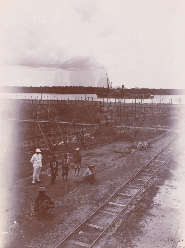 Overview of part of the construction pit and 9th caisson, photograph, Construction site with supervisor and workers, a track on the right. Numbered: 14. Part of the group of loose photographs regarding the construction of the port of Belawan on the coast off Medan, Sumatra 1921-1922., inscription: 'No. 14. Overview of part of the construction pit and 9th caisson', date: '2, 4-21', Belawan, 1921-04-02, photographer, Sumatra, paper, daylight gelatin silver print, height 220 mm x width 160 mm, harbour, building-site, railway, tramway, rack railway, economic history, overseas history, traffic and transport, infrastructure, harbor, Period, 1921 - 1922, A riverside scene with wooden scaffolding and walkways extending along the water, a large boat moored near the far bank, a stretch of rails running along the muddy foreground, several men standing and sitting on the ground wearing hats and light clothing, a pile of goods or materials near the shore, and a distant treeline under a cloudy sky.