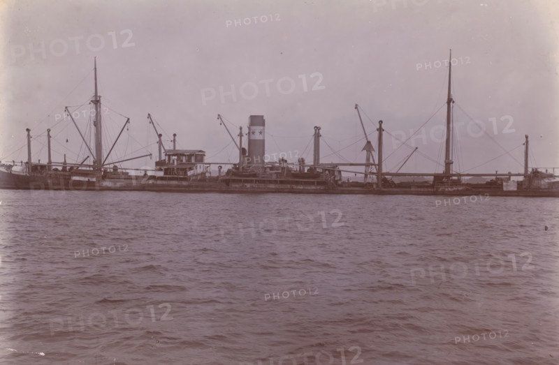 Image of SS Altenburg in Singapore, Belawan Ocean Harbor, photograph, The ship SS Altenburg in Singapore. Numbered: 71. Part of the group of individual photographs regarding the construction of the port of Belawan on the coast off Medan, Sumatra 1921-1922., inscription: 'No. 71, Image of SS Altenburg in Singapore', date: '23, 9-21', Singapore, 1921-09-23, photographer, paper, daylight gelatin silver print, height 160 mm x width 220 mm, steamsailer, economic history, overseas history, traffic and transport, infrastructure, harbor, Sumatra, Belawan, Period, 1921 - 1922, A long steamship viewed from the water with a tall smokestack near the center, multiple masts and booms connected by rigging, a raised wheelhouse and deck structures amidships, lifeboats and cargo gear visible along the deck, calm rippling water in the foreground, and a pale sky above.