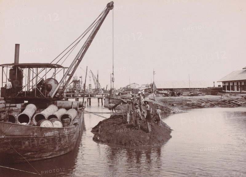 Closing of the dam around the construction pit, Belawan Ocean Harbor, photograph, Closing the dam around the construction pit. Numbered: 102. Part of the group of individual photographs regarding the construction of the port of Belawan on the coast off Medan, Sumatra 1921-1922., inscription: 'No. 102 Closing of the dam around the construction pit', date: '20, 12-21', Belawan, 1921-12-20, photographer, Sumatra, paper, daylight gelatin silver print, height 160 mm x width 220 mm, harbor, building-site, economic history, overseas history, traffic and transport, infrastructure, dredging, Period, 1921 - 1922, A riverside worksite shows a large crane mounted on a barge with a tall boom and cables, a winch and a covered engine area, and several large cylindrical pipes lying on the deck of the barge; a suspended bucket hangs over the water while workers wearing hats and light clothing stand and work on a muddy embankment beside the water, some holding shovels; in the background are wooden piers...