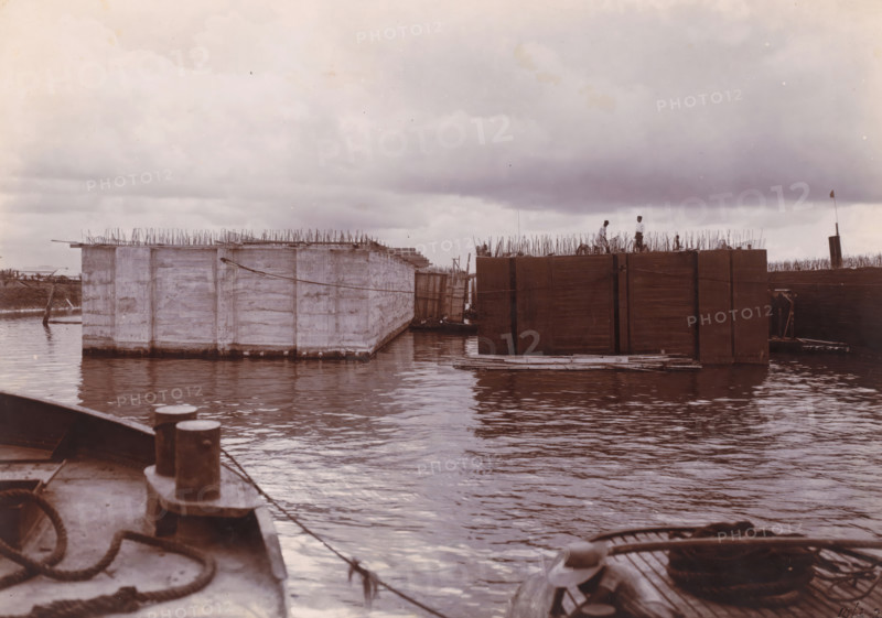 Dismantling, plastering and tarring of the 9.09 m high caissons. (1st work), Ocean Harbor Belawan, photograph, Finishing of the caissons. Numbered: 129. Part of the group of loose photographs about the construction of the harbor Belawan on the coast off Medan, Sumatra 1921-1922., date: '10, 3-22', inscription: '129 Dismantling, plastering and tarring of the 9.09 m high caissons. (1st work)', Belawan, 1922-03-10, photographer, Sumatra, paper, daylight gelatin silver print, height 160 mm x width 220 mm, harbor, building-site, economic history, overseas history, traffic and transport, infrastructure, dredging, Period, 1921 - 1922, A waterscape with large rectangular floating structures framed by wooden and concrete surfaces, vertical metal rods protruding from their tops, two people standing on one of the structures, calm rippling water reflecting the sky, a foreground view of a vessel deck with mooring posts, ropes, and a coil, a small boat tied alongside one of the floating platforms, d...