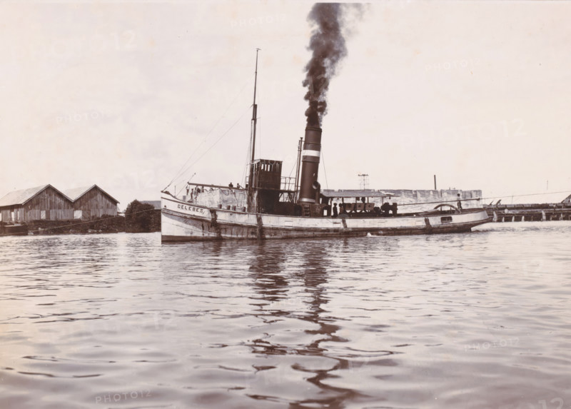 Tugboat 'Celebes', Ocean Harbor Belawan, photograph, The tugboat 'Celebes' with smoking chimney. Numbered: 163. Part of the group of individual photographs regarding the construction of the harbor of Belawan on the coast off Medan, Sumatra 1921-1922., date: '25, 5-22', inscription: 'No 163 Tugboat 'Celebes'', Belawan, 1922-05-25, photographer, Sumatra, paper, daylight gelatin silver print, height 160 mm x width 220 mm, harbor, building-site, tug, pusher tug, economic history, overseas history, traffic and transport, infrastructure, dredging, Period, 1921 - 1922, A steamship with a tall smokestack emitting dark smoke is floating on calm water, its hull showing signs of wear and weathering and a small raised wheelhouse amidships, with several people visible on the deck near railings; a mast with rigging rises above the deck toward the bow, and the stern has a rounded cabin area. Along the shoreline behind the ship there are wooden buildings with pitched roofs, a low pier structure, and s...