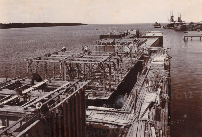 Panorama from construction jetty 5 caissons 1st phase fully cast, Ocean Harbor Belawan, photograph, View of the work on the caissons, at a jetty in the harbor, June 1922. Part of the group of loose photographs of the construction of the harbor of Belawan on the coast off Medan, Sumatra 1921-1922., date: '17, 6-22', inscription: 'No. 170, Panorama from construction platform 5 caissons 1st work fully cast', Belawan, 1922-06-17, photographer, Sumatra, paper, daylight gelatin silver print, height 160 mm x width 220 mm, harbor, building-site, economic history, overseas history, traffic and transport, infrastructure, Period, 1921 - 1922, A wide waterfront scene shows an extensive wooden pier or dock under construction extending into calm water, with a complex framework of beams, planks, and scaffolding forming multiple rectangular sections. Several workers in plain clothing and hats are positioned on the wooden structures, some crouching or kneeling on beams and others standing on walkways b...