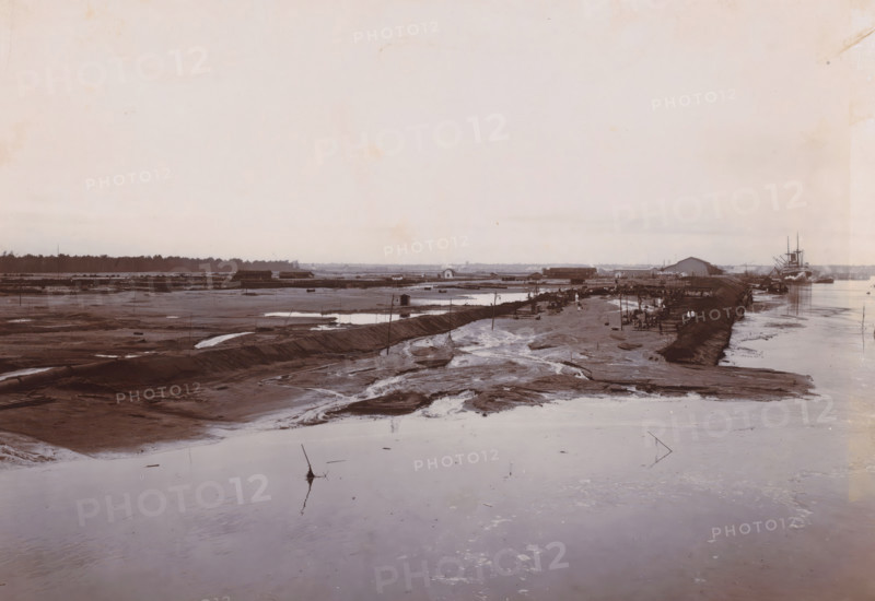 Overview of sand loading, Belawan Ocean Port, photograph, View of the reclaimed land. Numbered: 180. Part of the group of individual photographs regarding the construction of the port of Belawan on the coast off Medan, Sumatra 1921-1922., annotation: 'No. 180 Overview of sand loading', date: '14, 7-22', Belawan, 1922-07-14, photographer, Sumatra, paper, daylight gelatin silver print, height 160 mm x width 220 mm, harbour, building-site, economic history, overseas history, traffic and transport, infrastructure, harbor, dredging, Period, 1921 - 1922, A wide, muddy riverscape fills the scene with shallow pools and meandering wet channels in the foreground, a raised muddy embankment running diagonally toward the right where a cluster of small wooden structures and groups of people are gathered along the water's edge, nearby boats and a larger steamship moored at the far right with visible masts and smokestack, low buildings and warehouses stretch along the midground behind the embankment, ...
