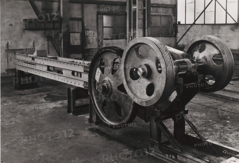 Ampas press, photograph, intended for an unknown sugar factory. Part of the binder containing photographs of machines from NV Machinefabriek Braat in Surabaya intended for the Dutch sugar industry in Java, from the period 1917-1938., Surabaya, 1917 - 1938, photographer, Java, East Java, paper, gelatin silver print, gelatin silver print on photographic paper, height 124 mm x width 180 mm, Person, NV Machinefabriek Braat, colonial system, (non-fruit) products of plants or trees: sugarcane, factory-interior, machine shop, machines, parts of machines, tools and appliances, economic history, overseas history, sugar industry, business history, Period, A large industrial machine dominates the foreground with two massive metal wheels featuring circular cutouts and grooved rims mounted on a heavy base, connected to a toothed gear and a cylindrical component, all showing worn and oily surfaces. A long metal framework with riveted beams and rectangular openings extends from the machine toward the...