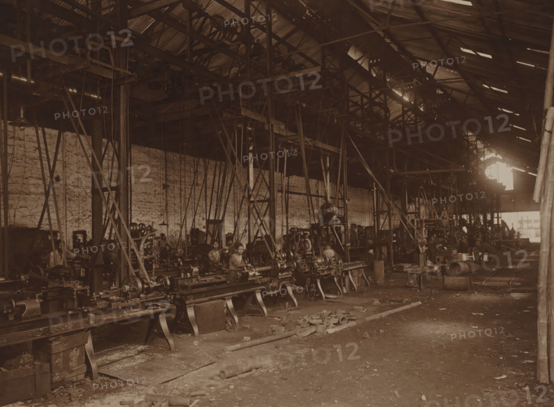 First turning shop, photograph, View into the factory hall with the first turning shop installed in 1904. Pictured here after the electrification in 1908. Part of the binder with photos of machines from NV Machinefabriek Braat in Surabaya, Sukabumi, and Yogyakarta, from the period 1908-1942., Surabaya, 1908, photographer, Java, East Java, paper, photography, height 153 mm x width 202 mm, Person, NV Machinefabriek Braat, colonial system, factory-interior, machine shop, machines, parts of machines, tools and appliances, economic history, overseas history, business history, Period, 1908 - 1942, A large industrial interior with rows of heavy metal lathes and machinery arranged on raised supports, a high corrugated metal roof with exposed beams and pulley systems, vertical and diagonal metal supports and belts hanging down toward the machines, a long brick wall along one side, several workers seated or standing at the machines, scattered metal parts and debris on the concrete floor, shafts ...