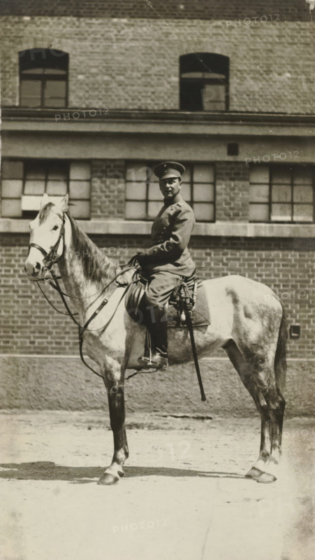 Arthur Moos on horseback in the uniform of the 20th Uhlanen regiment, May 1916, in Ludwigsburg, photograph, Arthur Moos on horseback in the uniform of the 20th Uhlanen regiment in Ludwigsburg., Ludwigsburg, 1916-05, photographer, Baden-Württemberg, paper, gelatin silver print, developing gelatin silver print, height 135 mm x width 80 mm, Person, Arthur Moos, Period, A black-and-white photograph showing a person in a uniform and cap seated on a light-colored horse in front of a brick building with multi-pane windows; the rider faces slightly toward the camera and holds the reins, wearing boots and a belted coat with visible buttons, and a sword or saber hangs at the saddle; the horse has a bridle and saddle with straps and stands on a paved surface, while the building behind shows rows of bricks, window frames, and a horizontal ledge.