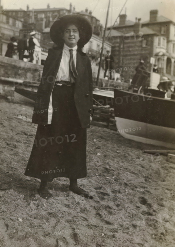 Else Wachenheimer-Moos with a hat in front of a boat on the beach, Bexhill-on-Sea, 1913, photograph, Bexhill-on-Sea., photographer, East Sussex, paper, gelatin silver print, developed gelatin silver print, height 110 mm x width 80 mm, Person, Else Wachenheimer-Moos, England, Period, A person stands on a sandy shore wearing a long dark skirt, dark jacket, light blouse with a scarf tied at the neck, and a wide-brimmed hat, with dark shoes visible on the sand; behind them a boat is beached with its hull partly visible and ropes and masts nearby, and further back there are buildings on a raised quay with several other figures and architectural details along the waterfront.