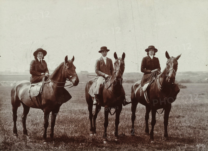 Else Wachenheimer-Moos and two unknown women on horseback, Bexhill-on-Sea in England, 1913, photograph, Else Wachenheimer-Moos (left) and two unknown women on horseback, Bexhill-on-Sea in England., Bexhill-on-Sea, photographer, East Sussex, paper, gelatin silver print, developing gelatin silver print, height 240 mm x width 310 mm, Person, Else Wachenheimer-Moos, England, Period, Three people wearing dark riding coats and wide-brimmed hats sit astride three dark horses in a grassy field; the riders hold reins and one holds a crop, the horses wear saddles and bridles and face forward with visible ears and manes, and the background shows a pale sky above a distant, low horizon.