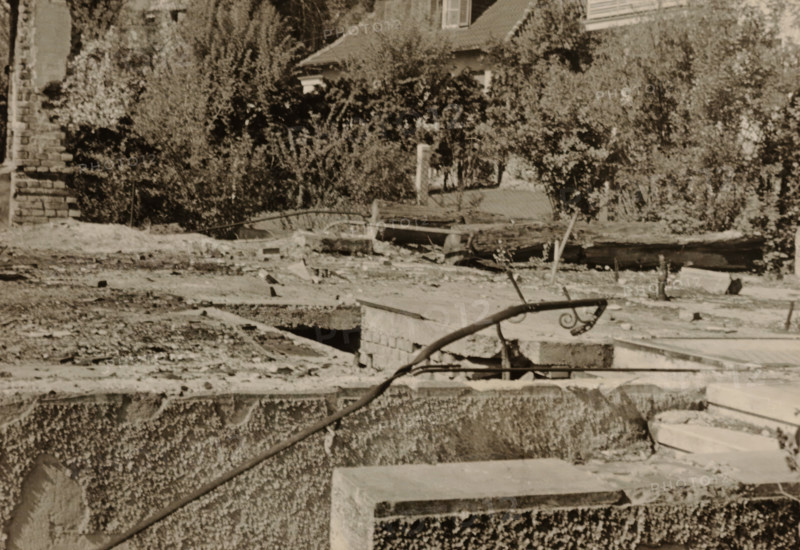 Top of part of a ruin, with a tree-lined house in the background, photograph, with a tree-lined house in the background., 1945 - 1970, photographer, paper, gelatin silver print, developed gelatin silver print, height 75 mm x width 105 mm, Period, A sepia-toned scene showing a rough concrete foreground with exposed edges and a curved metal handrail topped with a decorative spiral, rubble and broken slabs scattered across a flat midground with openings and gaps, a low stone wall and a rusted pipe lying across the surface, and dense shrubs and trees behind with partial views of a tiled roof and a window peeking through the foliage.