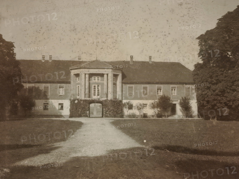 Front view of a country house with a gazebo-shaped central section, with trees on either side and a lawn and driveway at the front, photograph, with trees on either side and a lawn and driveway at the front., 1900 - 1950, photographer, paper, gelatin silver print, developed gelatin silver print, height 80 mm x width 105 mm, Period, A sepia-toned photograph of a large two-story building with a central portico supported by columns and an arched passage at ground level, flanked by windows on both sides and a pitched roof with chimneys; a gravel path leads from the foreground toward the archway, with lawns on either side and tall trees partially framing the building at the left and right edges of the image.