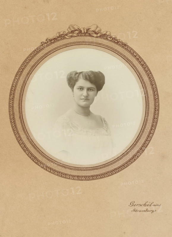Passepartout of Mathilde Wachenheimer-Wertheimer as a young woman, photograph, in light clothing., photographer: Gerschel aîné, Strasbourg (France), 1880 - 1895, paper, gelatin silver print, developed gelatin silver print, height 165 mm x width 105 mm, Person, Mathilde Wachenheimer-Wertheimer, Period, A sepia-toned portrait of a woman is centered within an ornate oval frame printed on textured paper; the woman faces forward with a neutral expression, her dark hair styled in a symmetrical updo, and she wears a light-colored dress with lace trim and a delicate necklace, while the decorative oval border features intricate patterned bands and a ribbon-like motif at the top.