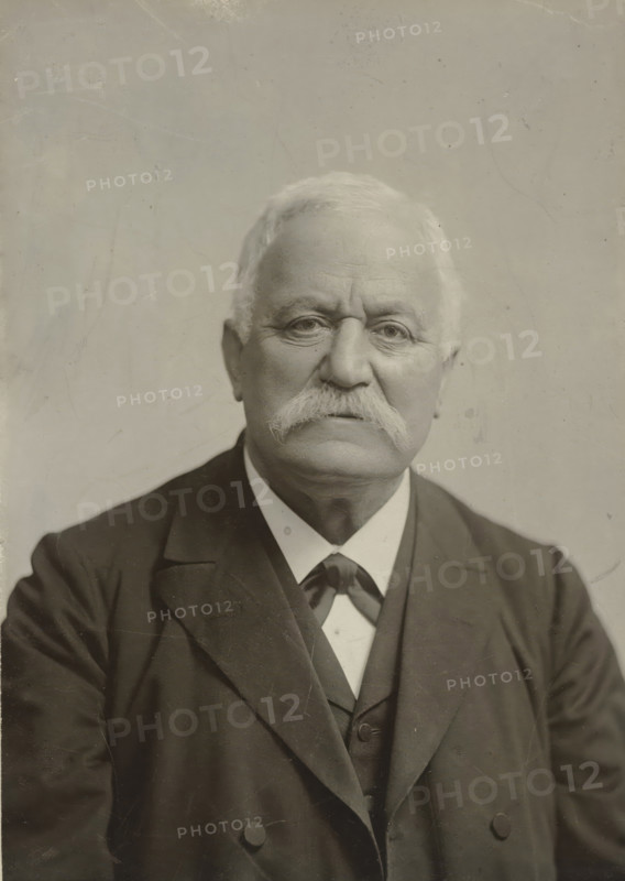 Portrait of an older man with a mustache, in dark clothing, photograph, in dark clothing., photographer: Gerschel aîné, Strasbourg (France), 1890 - 1898, paper, gelatin silver print, developed gelatin silver print, height 190 mm x width 140 mm, Period, 1890 - 1940, A close-up portrait of an older man with light hair and a thick white mustache, wearing a dark suit jacket over a waistcoat and a light-colored shirt with a necktie; his face is centered and shows visible skin texture and wrinkles, with neutral expression and eyes looking forward, and the background is a plain, slightly textured light surface.