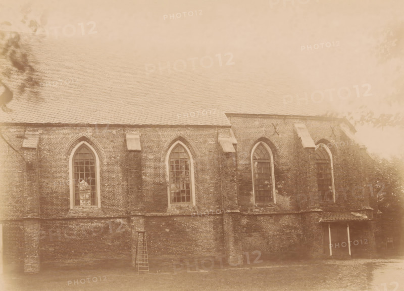 South side of the St. Nicholas Church in Dwingeloo, photograph, annotation: '12', annotation: 'Dwingelo', Dwingeloo, 1890 - 1910, photographer, Attributed to, paper, pasteboard, albumen print, height 166 mm x width 228 mm, church (exterior), Sint-Nicolaaskerk (Dwingeloo), A sepia-toned photograph of a brick building wall with a sloped tiled roof, showing a row of tall arched windows with multi-pane glazing set into the brickwork, buttresses between the window bays, small stone ledges above some sections, a wooden ladder leaning against the wall, tree branches and leaves partially visible at the image edges, and a ground area in front of the building.