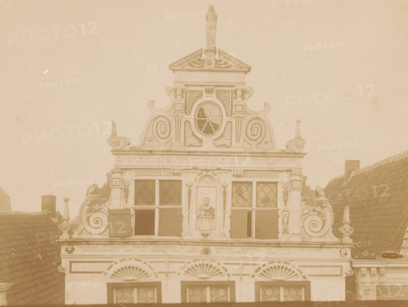 Facade of Friesestraat 9 in Coevorden, photograph, annotation: 'Friesesraat D.489, Coevorden', annotation: 'I', Coevorden, 1890 - 1910, photographer, Attributed to, paper, pasteboard, albumen print, albumendruk, height 168 mm x width 225 mm, façade (of house or building), An ornate building facade with decorative scrollwork and sculpted figures, a triangular pediment topped by a standing statue, an oval window with radial muntins centered beneath the pediment, several rectangular windows with diamond-pane glazing and open shutters, carved ornamental panels and leafy motifs around the openings, curved rooftop elements and finials on either side, a pair of stylized shell-shaped reliefs above lower windows, portions of sloped tiled roofs visible at the sides, and a muted sepia tone across the entire image.