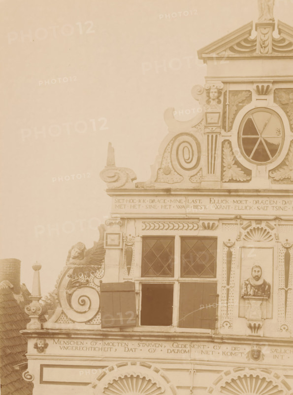 Facade of Friesestraat 9 in Coevorden, photograph, annotation: '18', annotation: 'Coevorden', Coevorden, 1890 - 1910, photographer, Attributed to, paper, pasteboard, albumen print, height 224 mm x width 168 mm, façade (of house or building), A sepia-toned photograph of an ornate building facade showing decorative stonework and carved ornaments concentrated on the upper portion, with a triangular pediment at the top, a round window with radial muntins set within a carved surround, scroll-shaped volutes, leaf and shell motifs, and a small sculpted face near the roofline; below are rectangular diamond-paned windows with wooden shutters, a carved bust set into a niche on the right, rows of horizontal friezes with carved borders, and ornamental finials and relief faces integrated into the masonry.