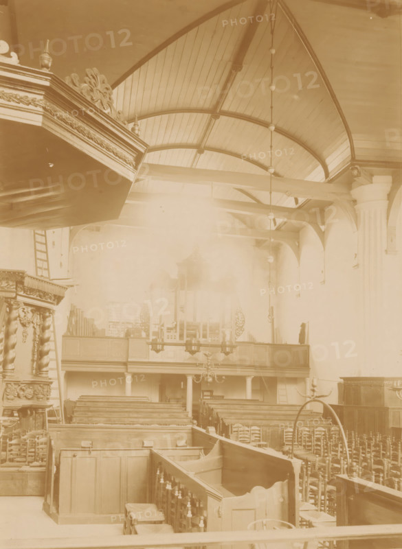 Interior of the Reformed Church in Coevorden, photograph, annotation: 'IV', annotation: 'Church in Coevorden', Coevorden, 1890 - 1910, photographer, Attributed to, paper, pasteboard, albumen print, height 229 mm x width 165 mm, interior of church, A sepia-toned interior view showing wooden pews arranged in rows, carved railings and paneling, ornate decorative woodwork on a raised pulpit with spiral columns, a balcony with wooden front and columns beneath it, a suspended chandelier and a vertical hanging chain of light fixtures, a vaulted wooden ceiling with curved beams, a tall fluted column against the right wall, and various chairs and wooden furniture scattered throughout the space.